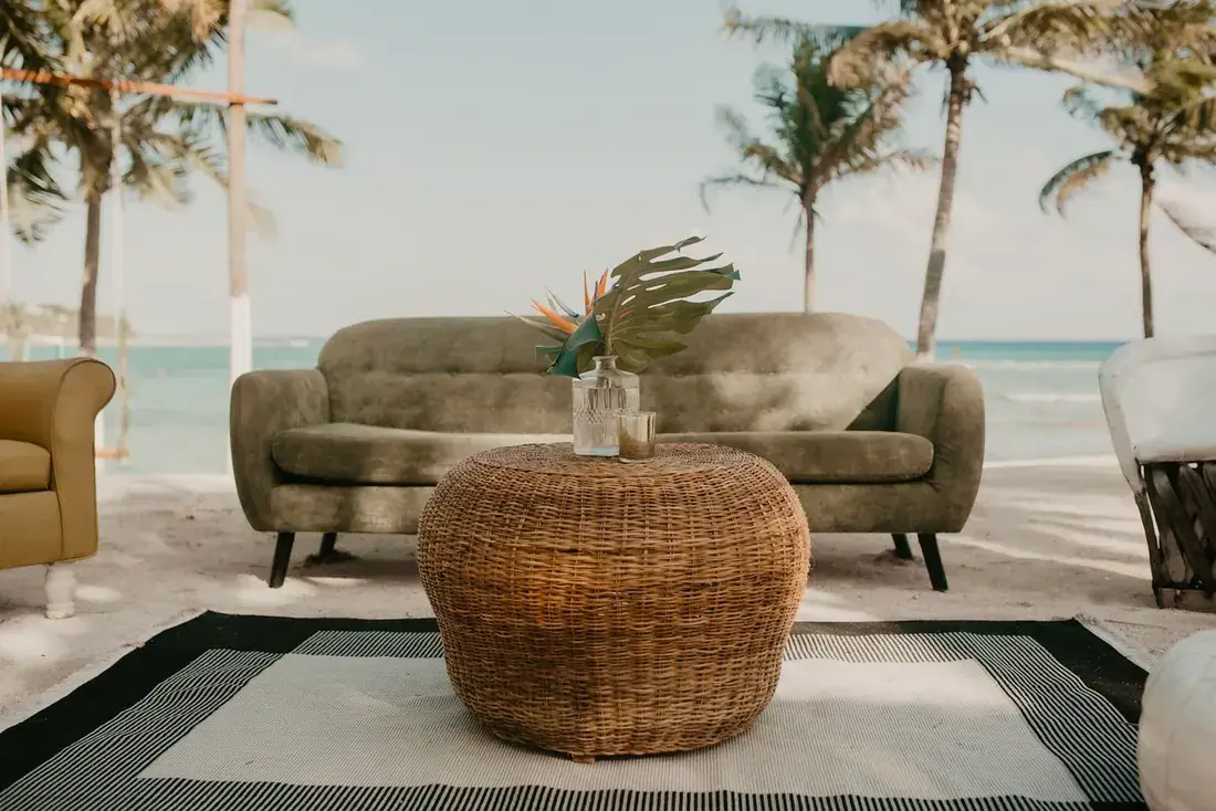 Green sofa and woven pouf on a rug on the beach beneath palms