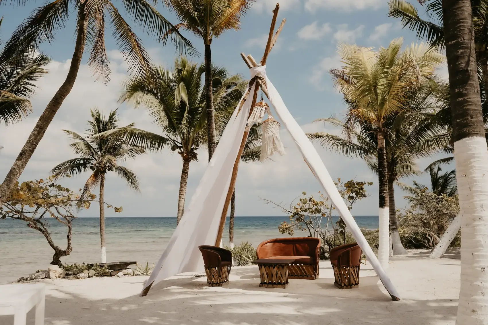 White teepee ceremony structure on the sand between palms, facing the sea