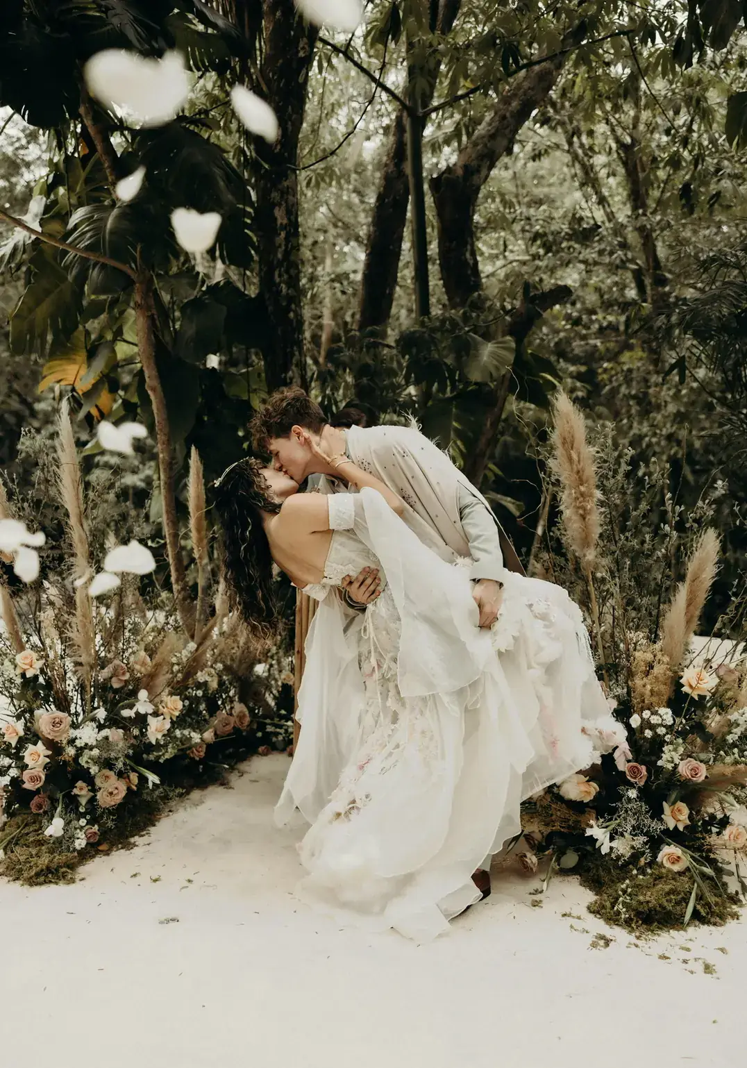 Bride in a flowing gown turning beside a jungle ceremony installation of white florals and dried palm