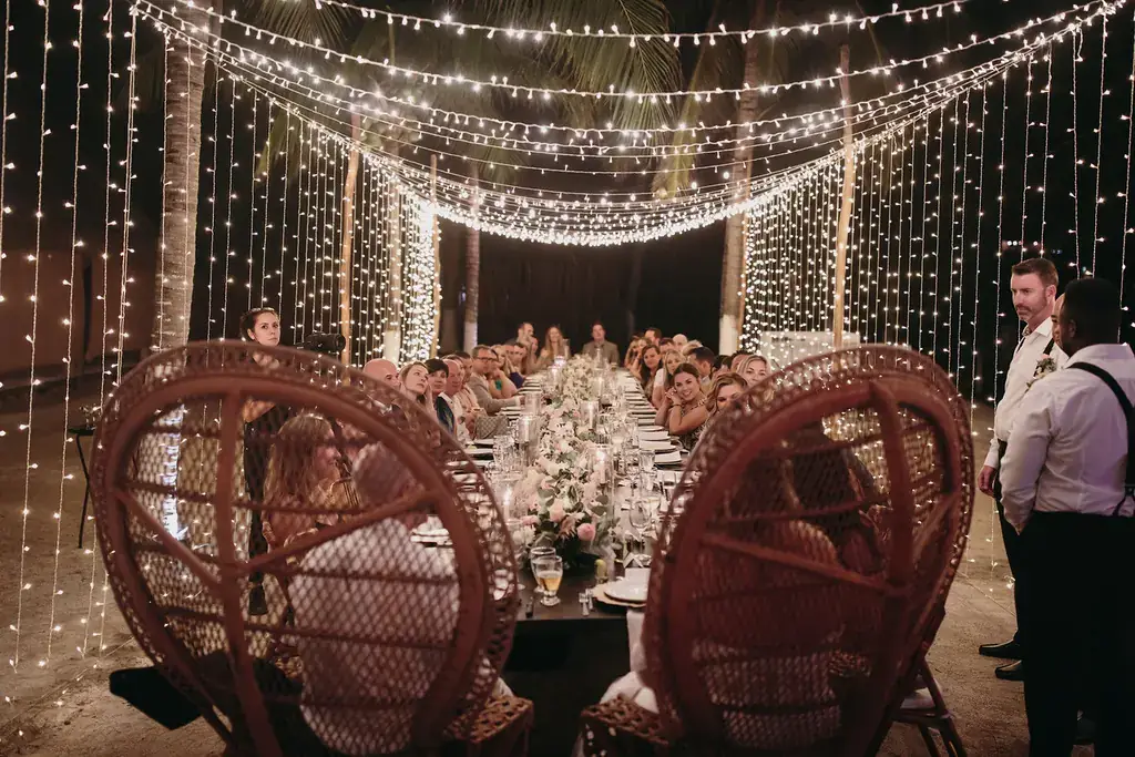 Long dinner table under a canopy of string lights and peacock chairs at night