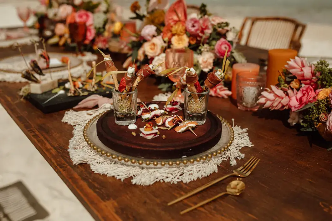 Moody wooden tablescape with coral and blush florals, gold cutlery, woven chargers and cut glassware