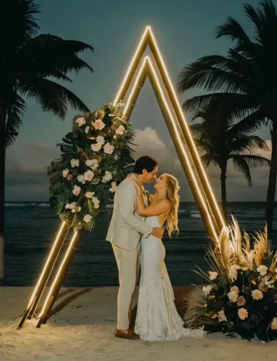 Couple embracing beneath an illuminated triangular arch dressed with florals on the beach at dusk