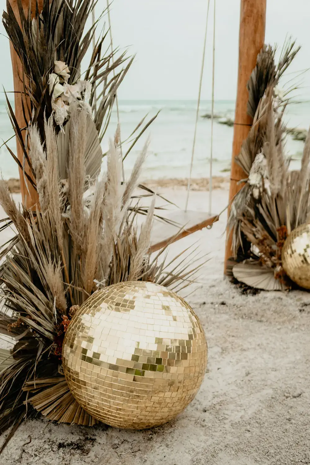 Gold mirror ball resting on sand between dried palm fronds