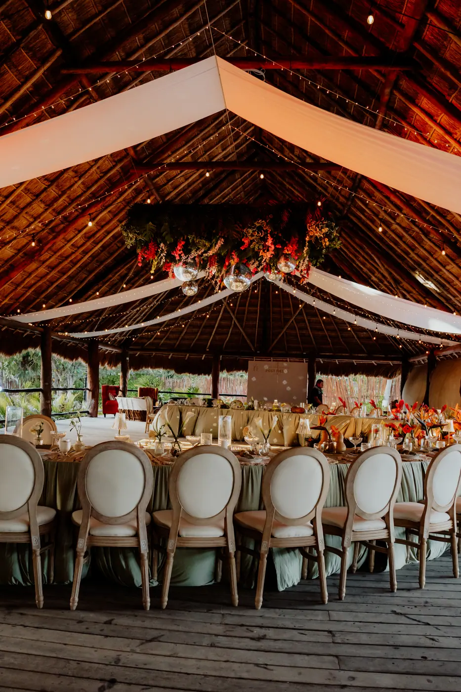 Palapa wedding reception with rattan chairs, olive draping and a hanging floral and disco ball installation